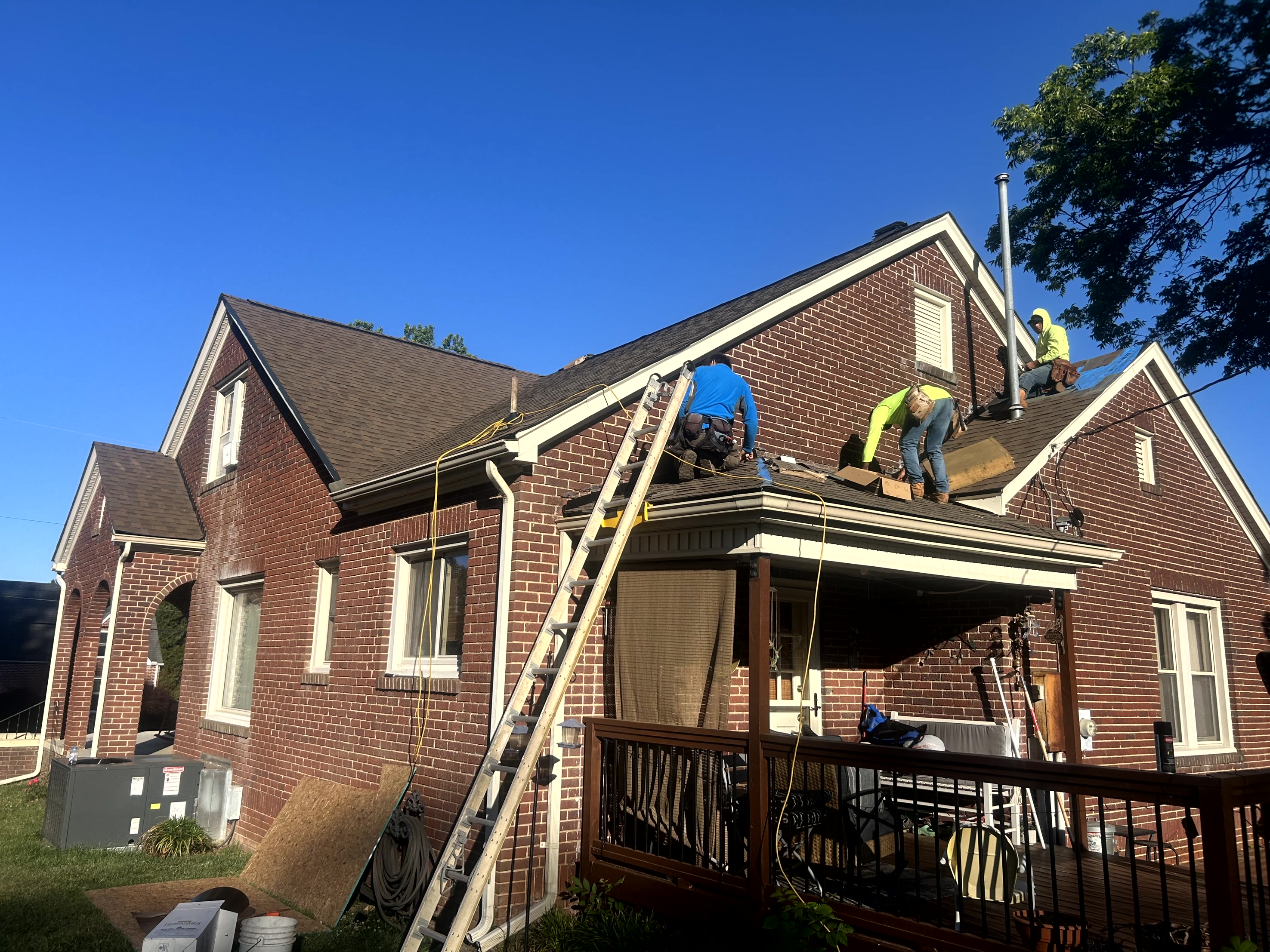 Three workers on a brick house roof repairing the shingles under a clear blue sky.