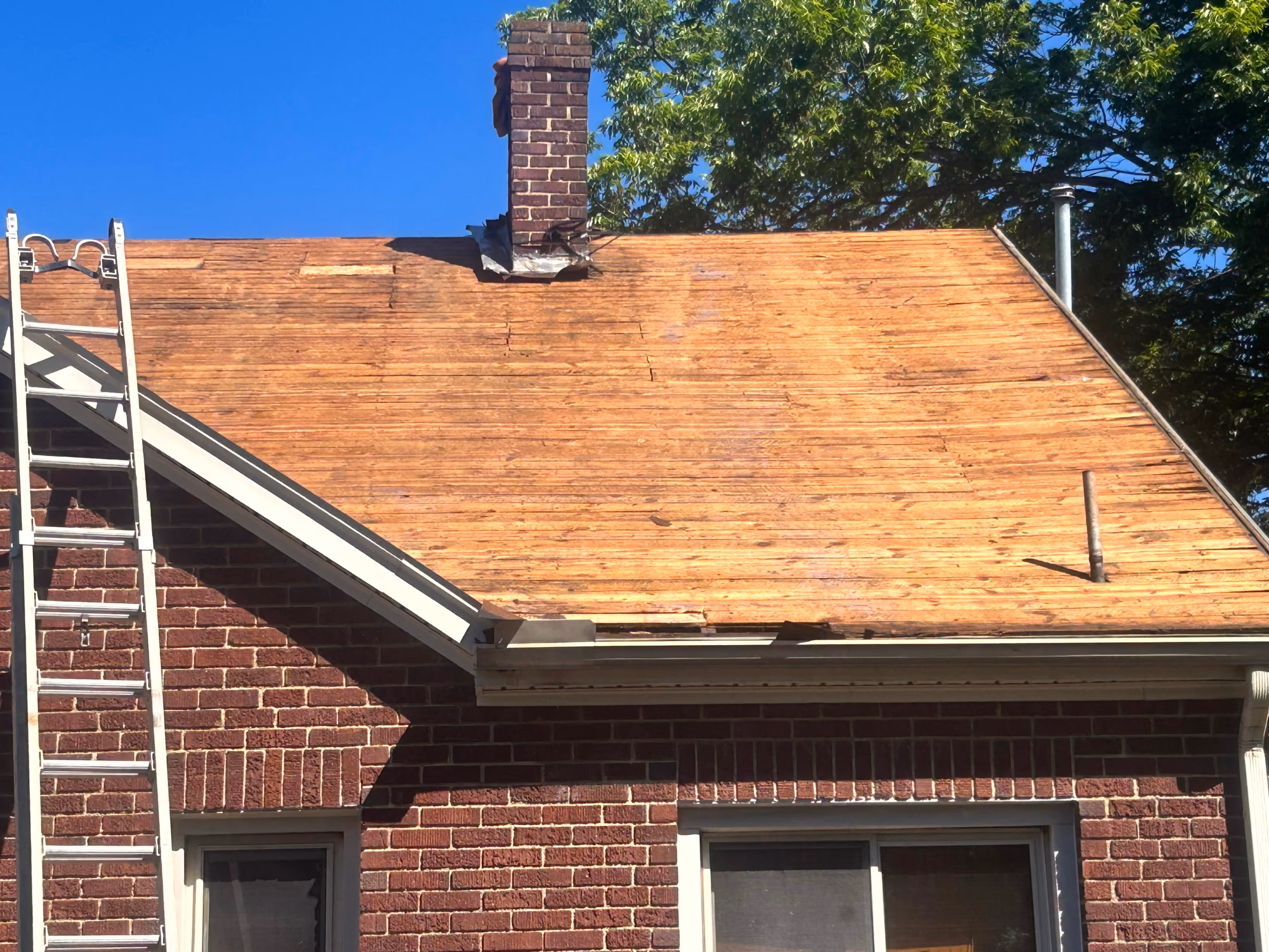 Partially stripped wooden roof of a brick house with a ladder leaning on the side under a clear blue sky.