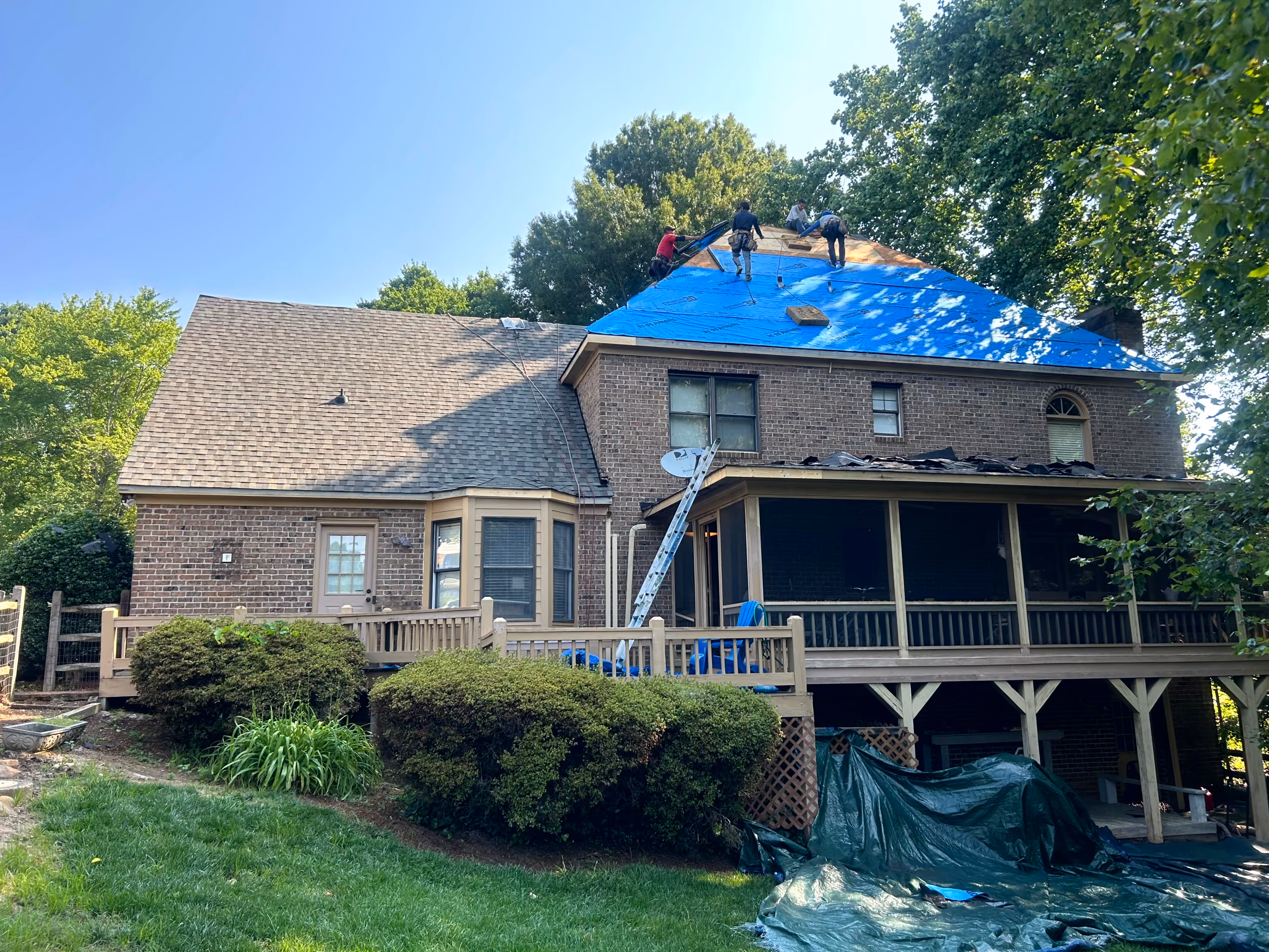 Workers repairing a large brick house roof covered partly with blue tarp under a clear sky.