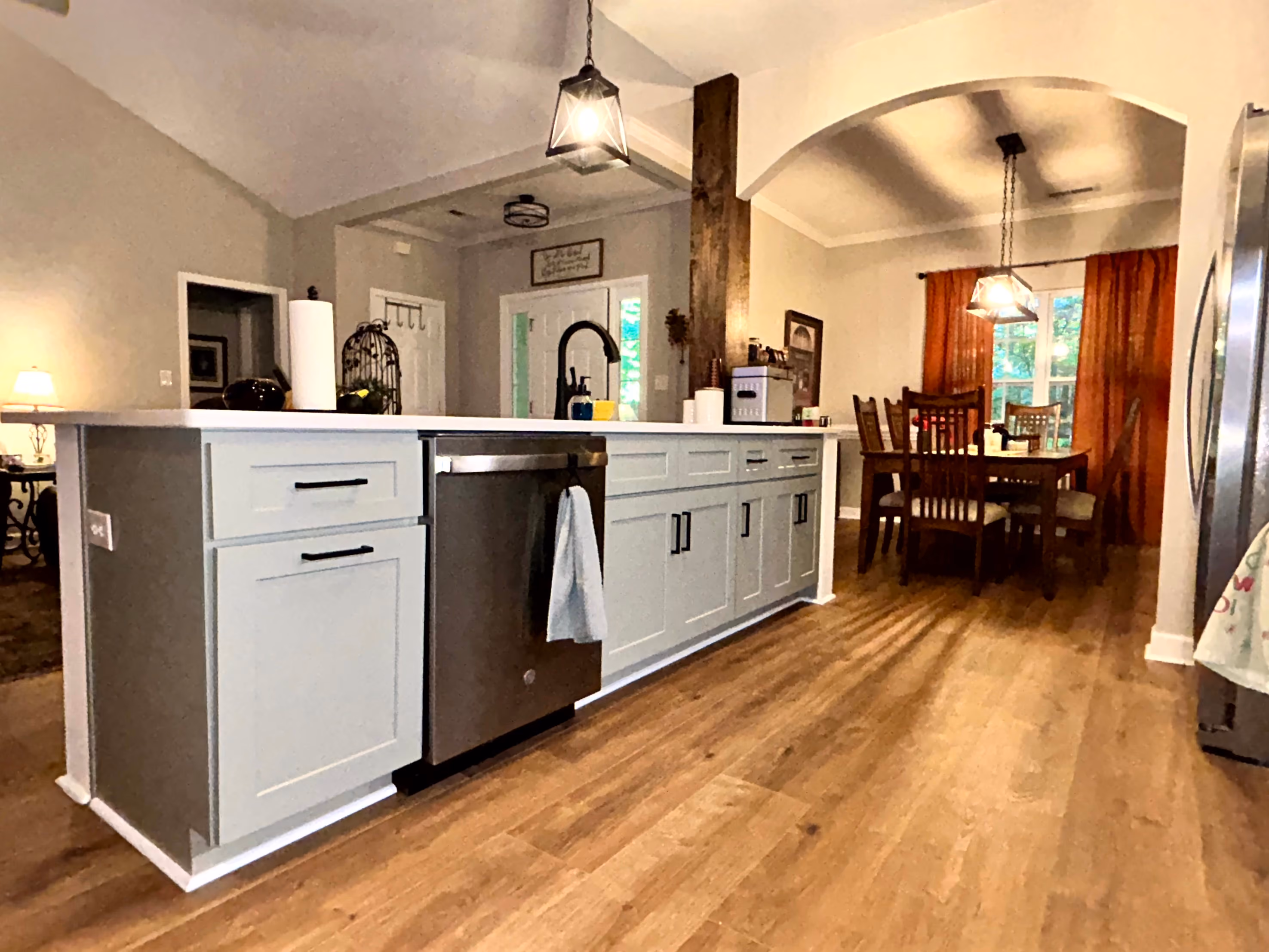 Modern kitchen island with white countertop, light gray cabinets, stainless dishwasher, dining area with wooden chairs.