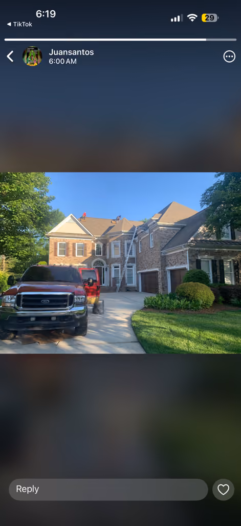 Two workers on a large house's roof with a ladder and parked red trucks in the driveway.