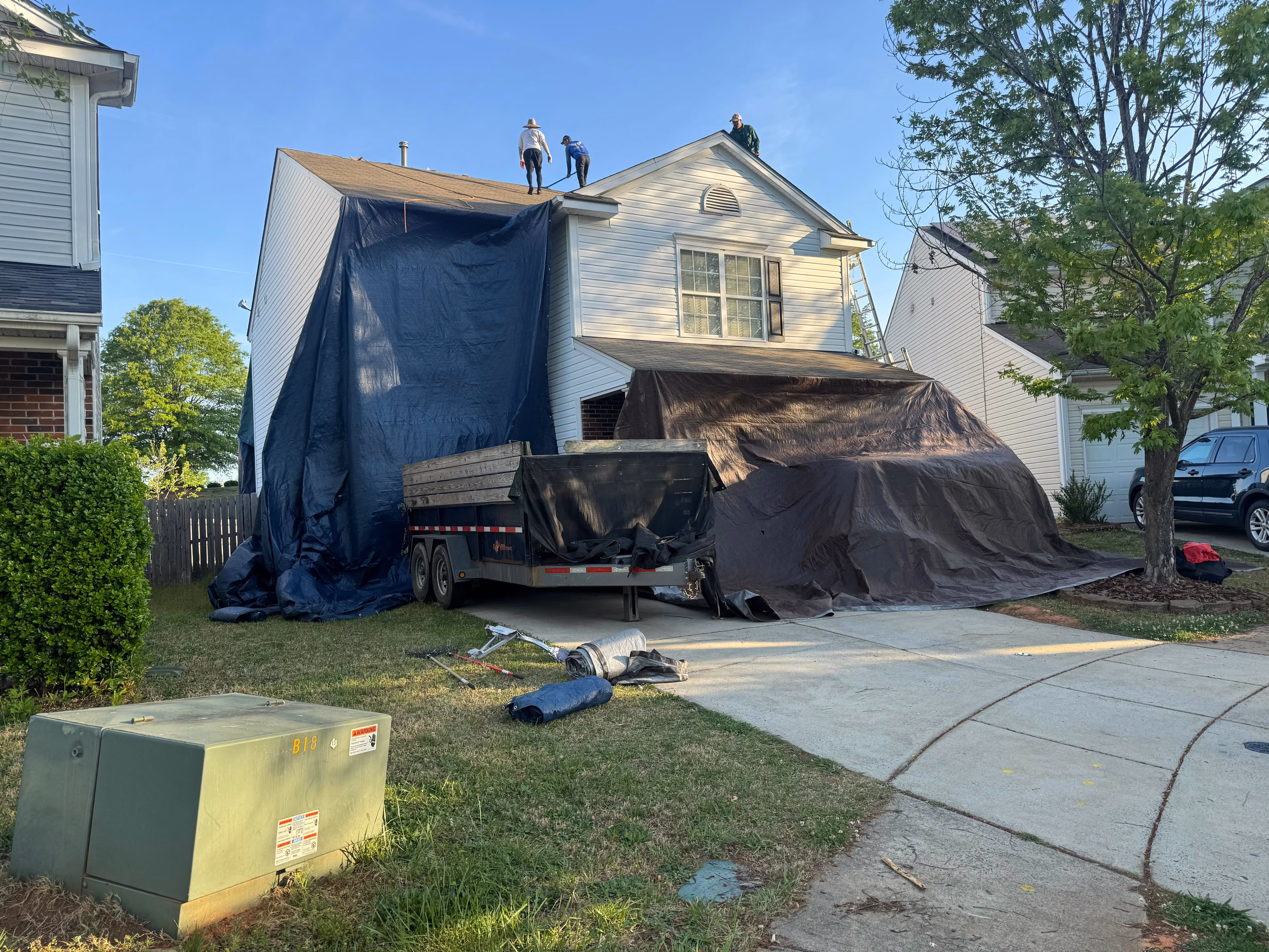 Three workers on a house roof covered with blue and brown tarps with tools and trailer in driveway.
