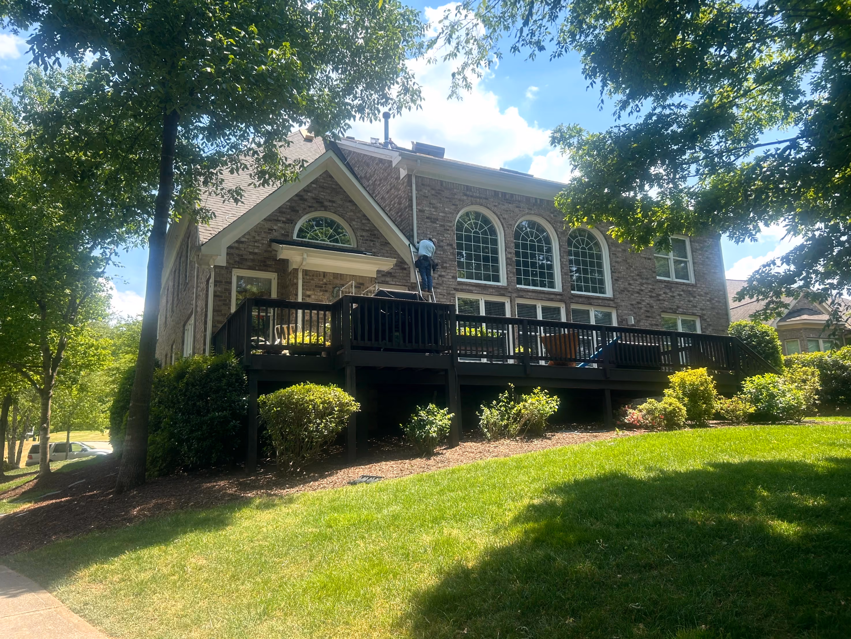 Brick house with large arched windows and wooden deck, man on ladder cleaning gutters.