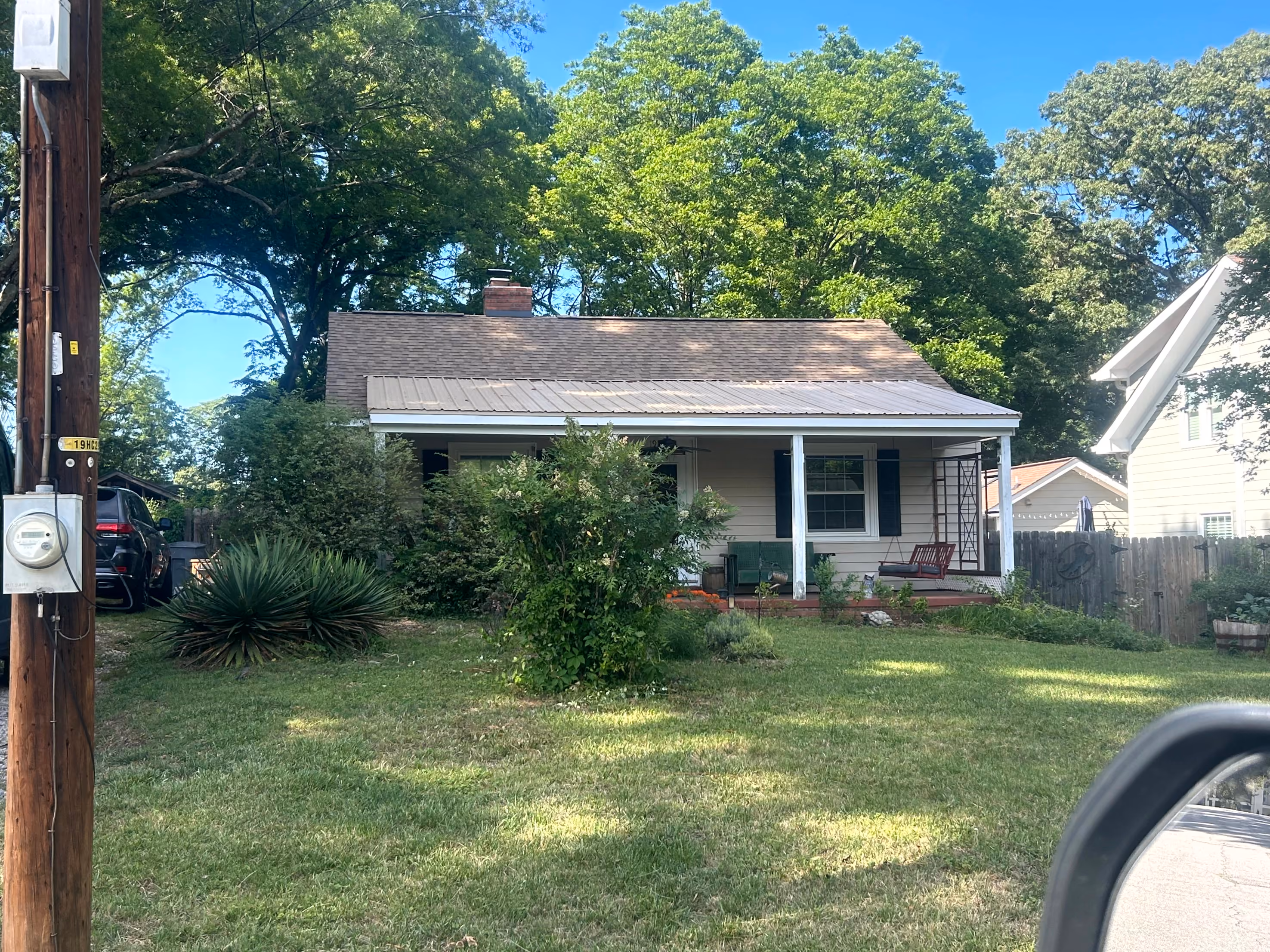 Small beige house with a front porch, surrounded by bushes, grass, and large trees under blue sky.