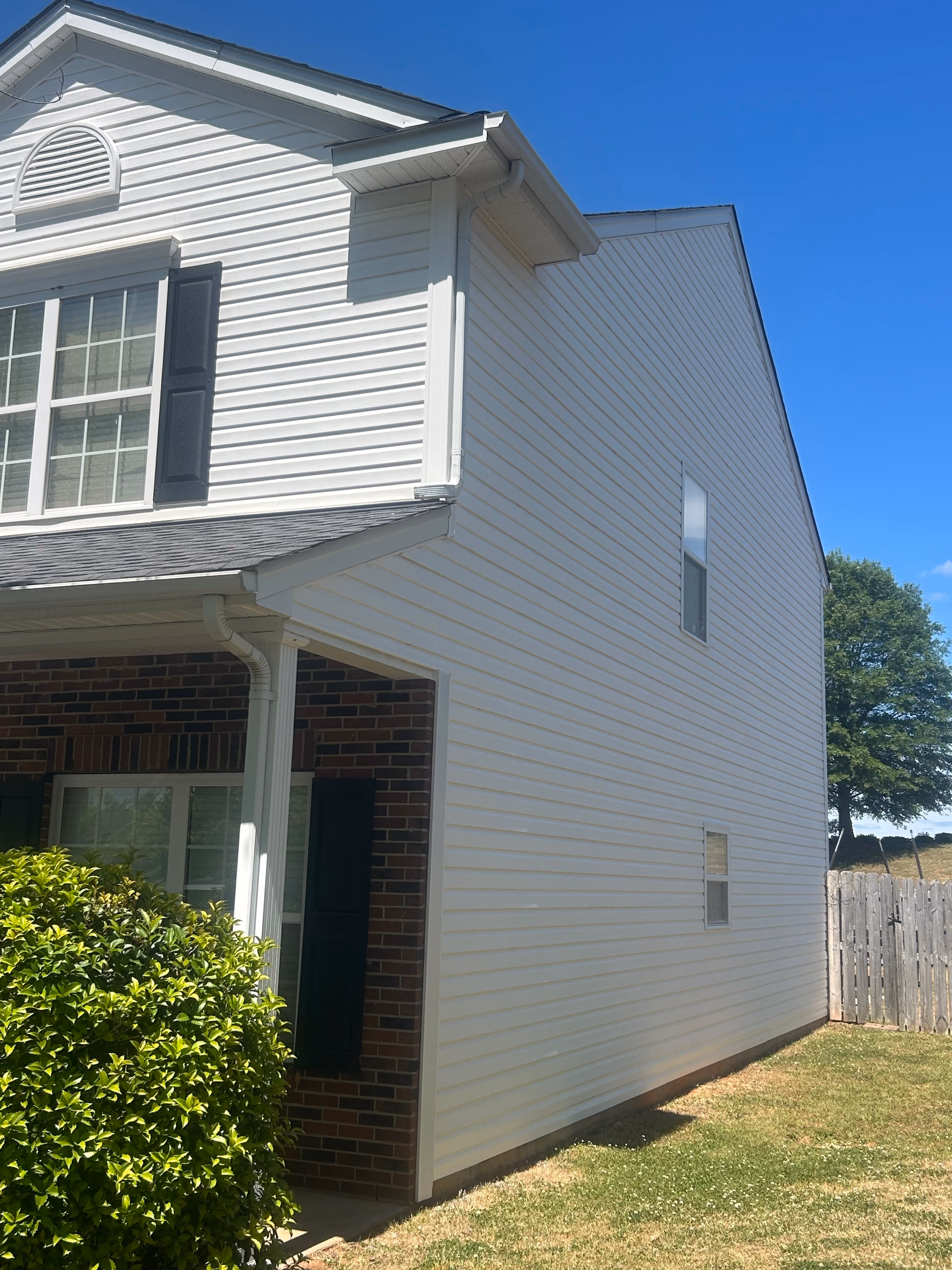 Side and front corner of a house with white siding, brick lower wall, two windows, and green shrub under blue sky.