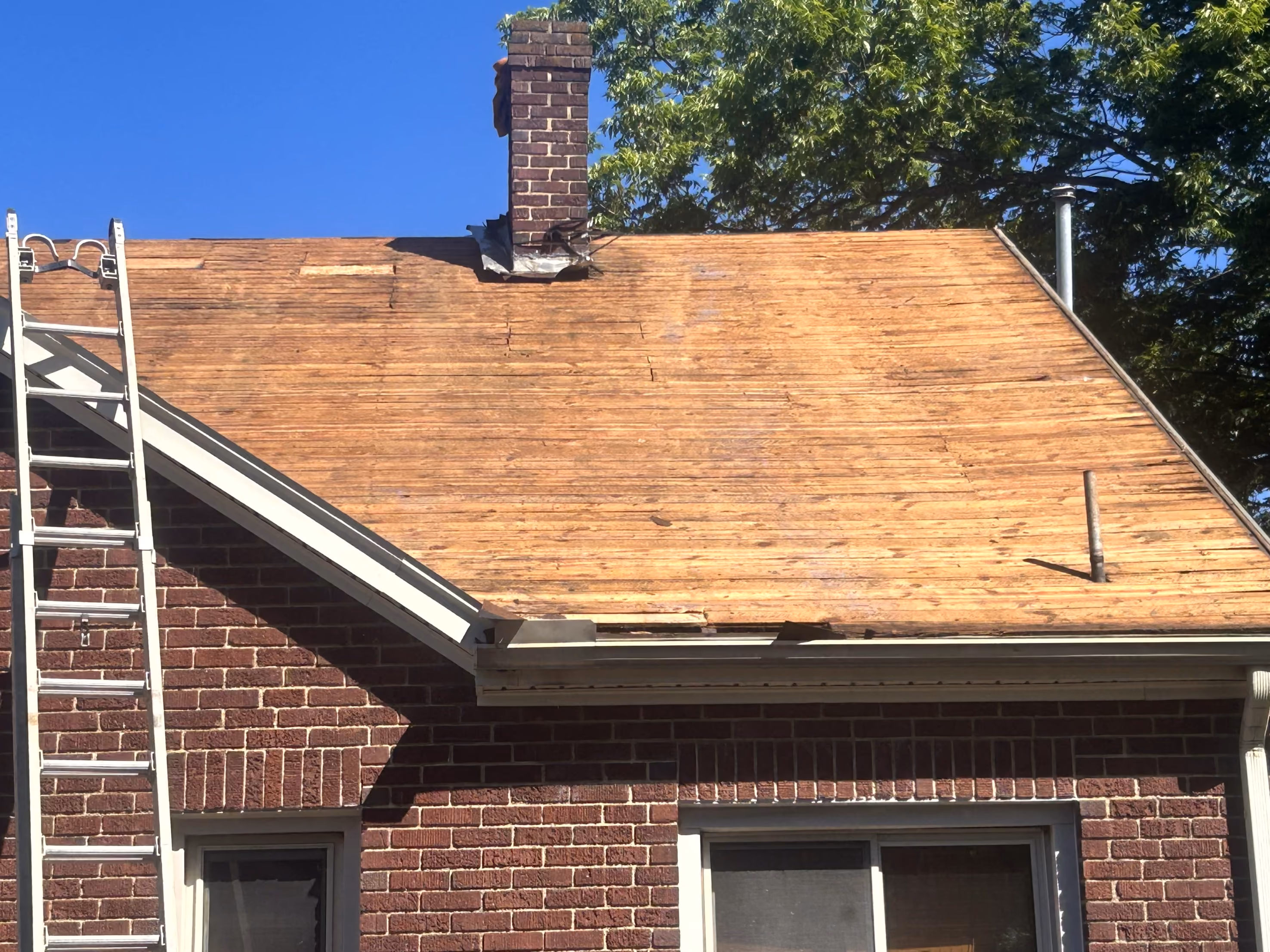 House roof stripped of shingles with chimney, ladder leaning on brick wall and tree in background.
