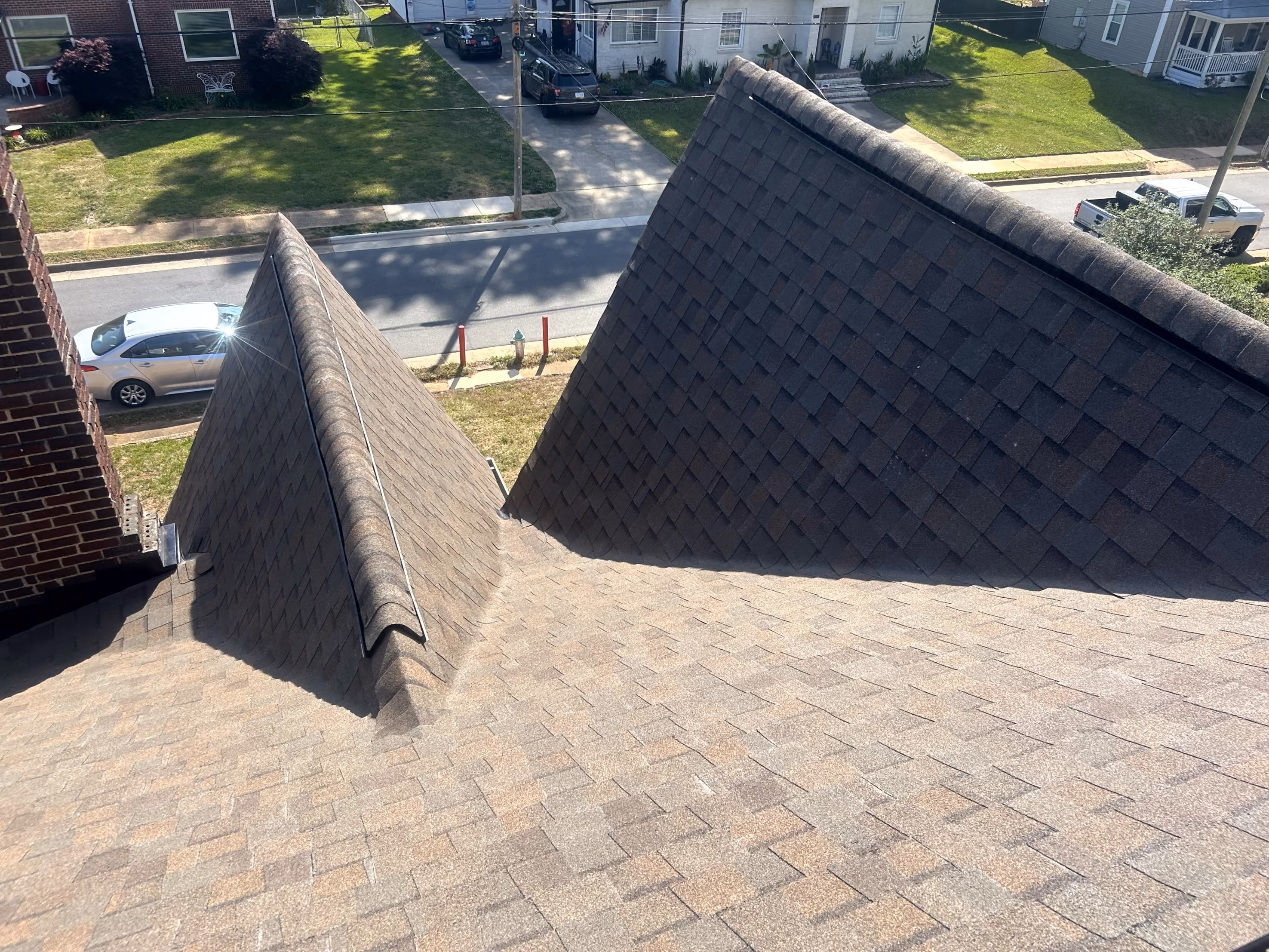 View of a roof with brown shingles and two peaked sections, with a street and parked cars in background.