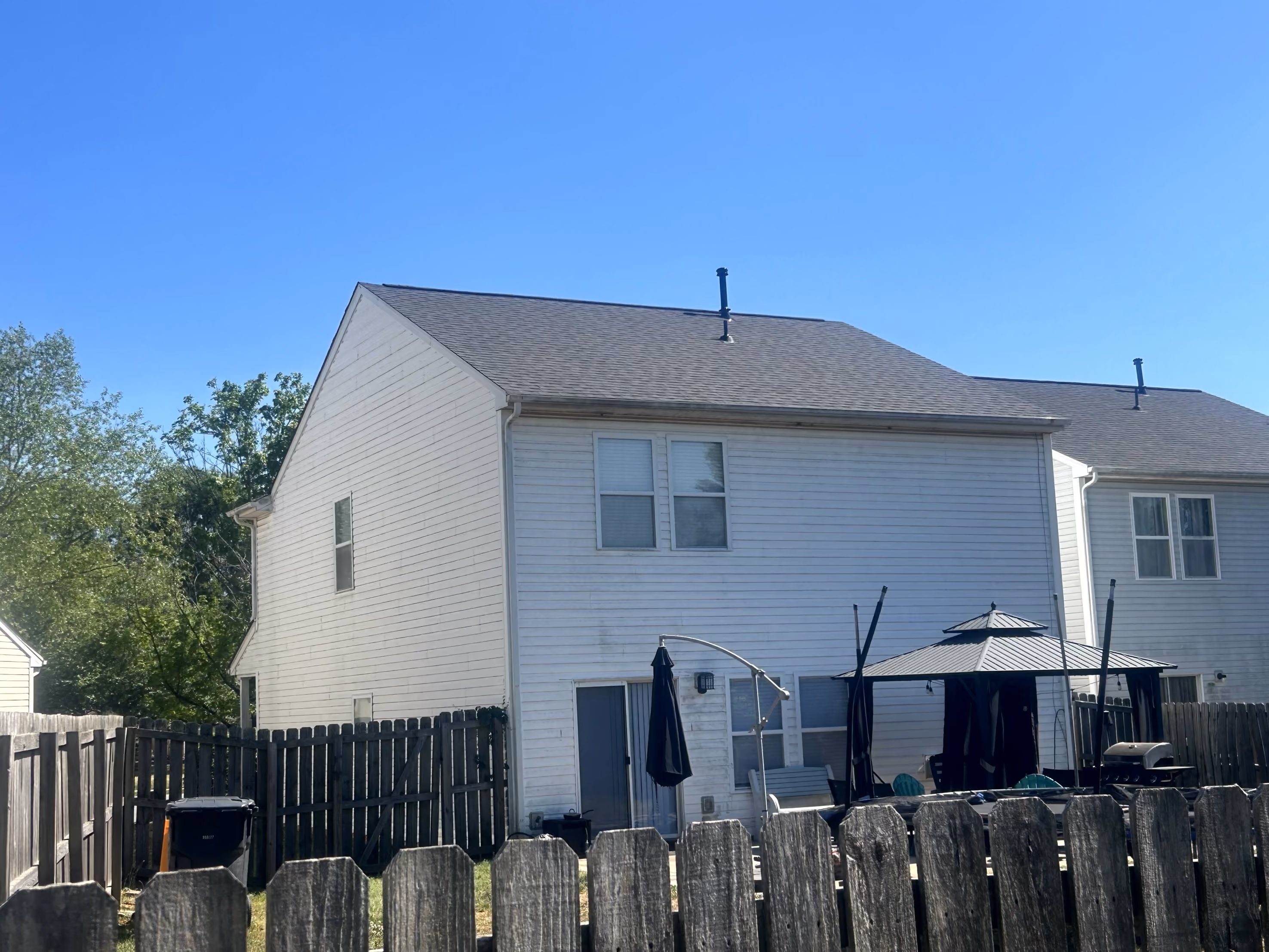 Backyard of two-story white house with wooden fence, patio umbrella, and grill on a clear day.