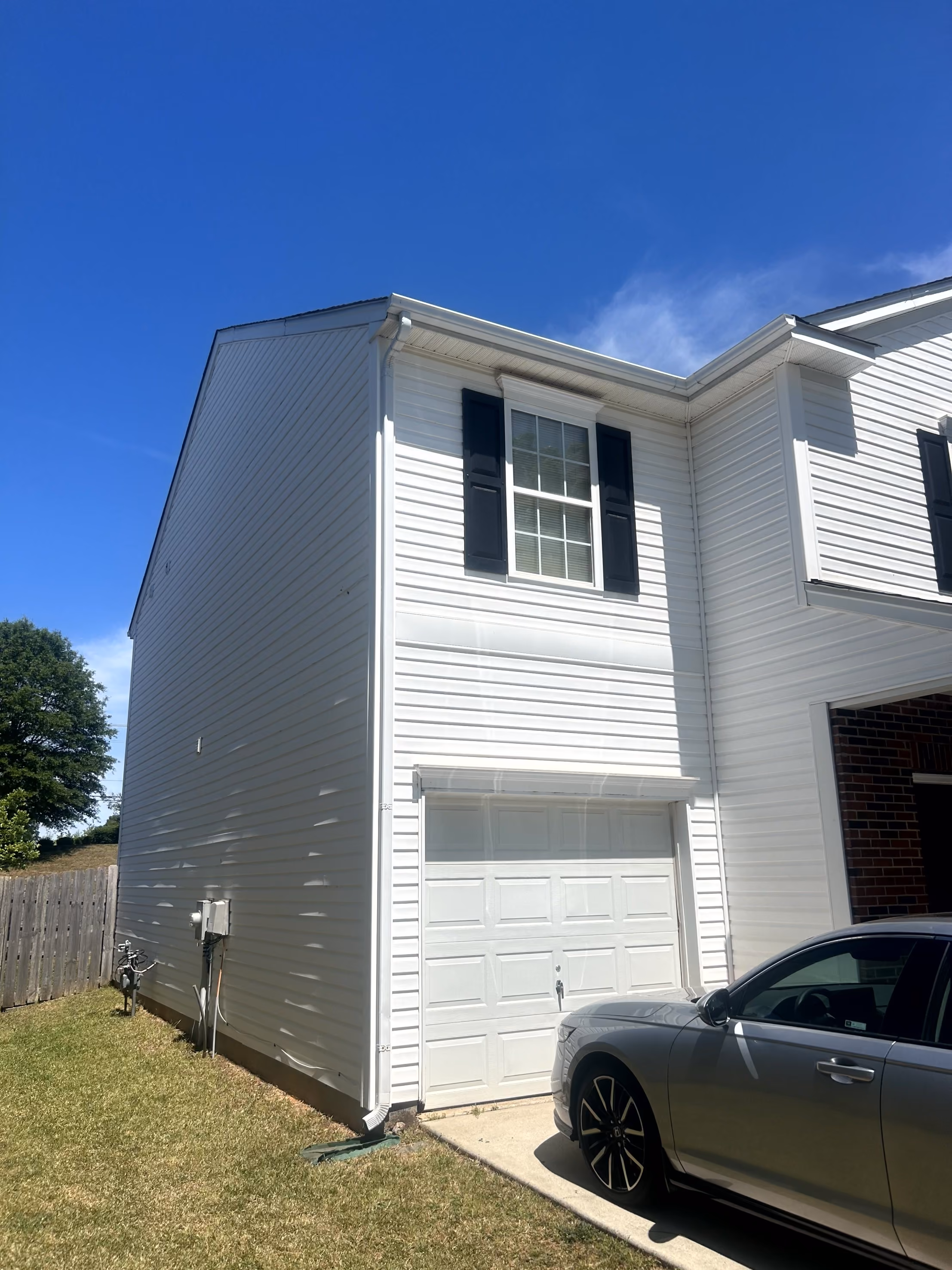 White two-story house side with garage door, window with black shutters, and silver car in driveway.