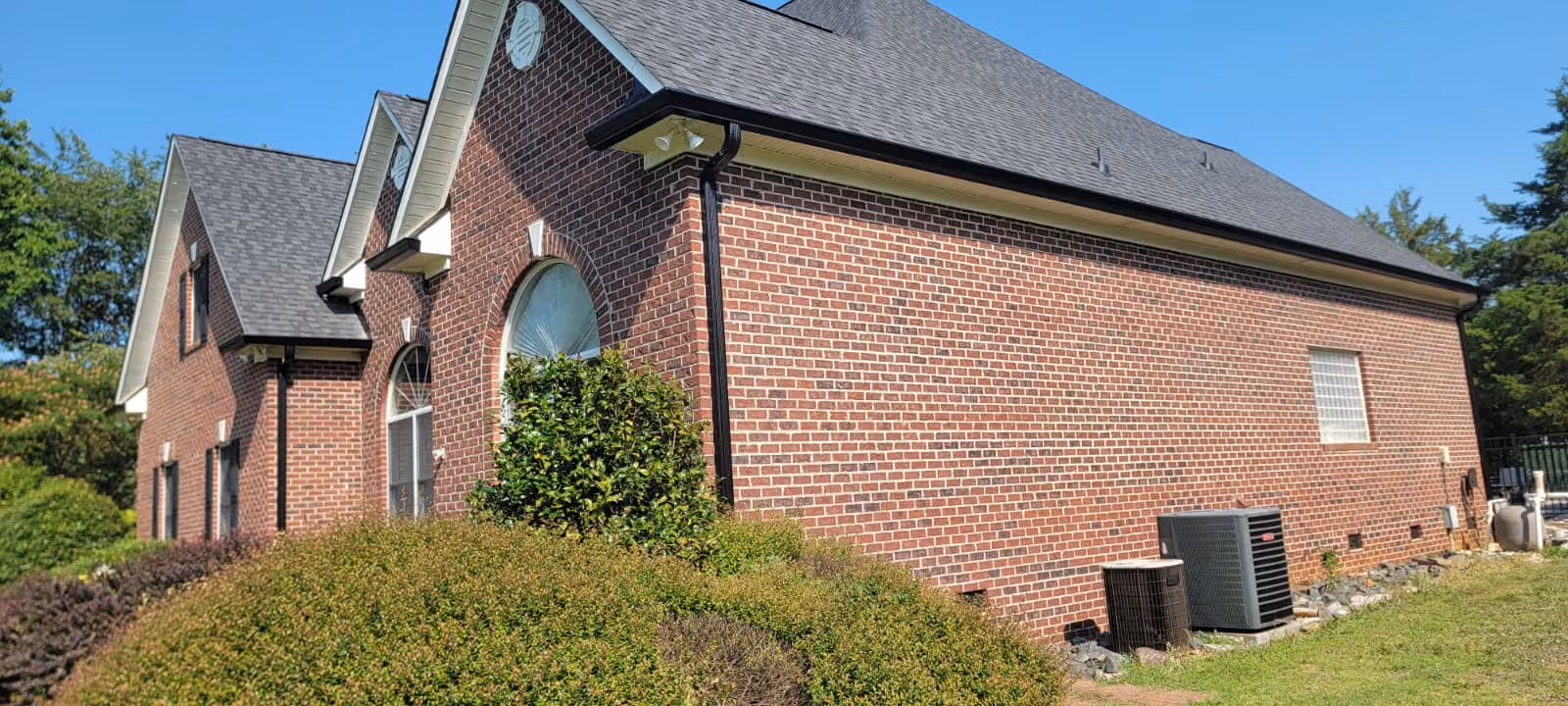 Side view of a brick house with arched windows, bushes, and two air conditioning units.