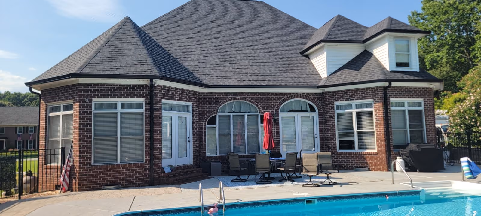 Brick house with dark gray roof, patio furniture, and a swimming pool in the foreground under clear sky.