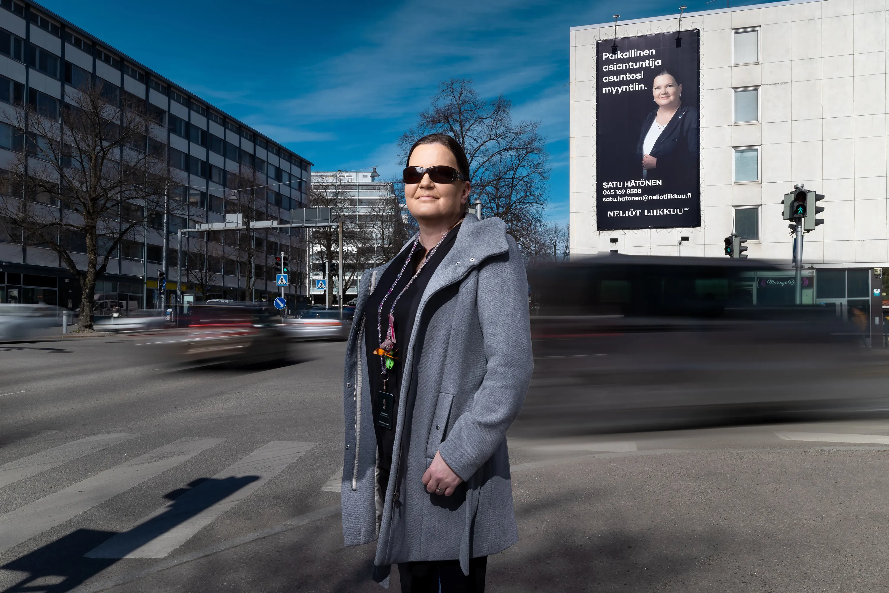 Aurinkolaseja ja harmaata takkia pitävä nainen seisoo Lahden kaupunkialueella kiinteistönvälittäjän mainostaulun edessä.