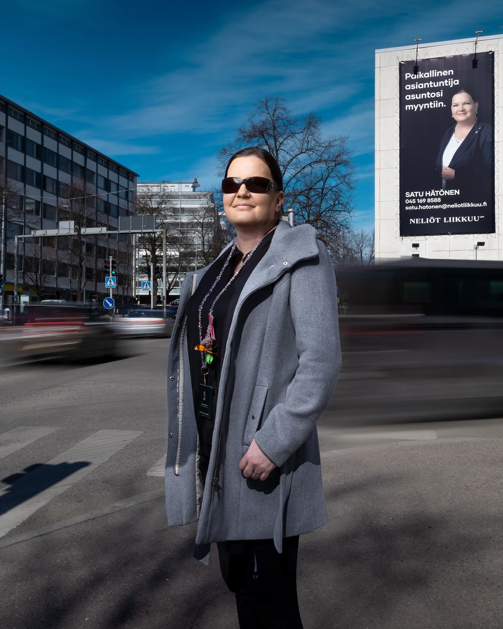 Nainen seisoo harmaassa takissa ja aurinkolaseissa kadulla, taustalla näkyy kaupunkimaisema ja suuri mainoskyltti.