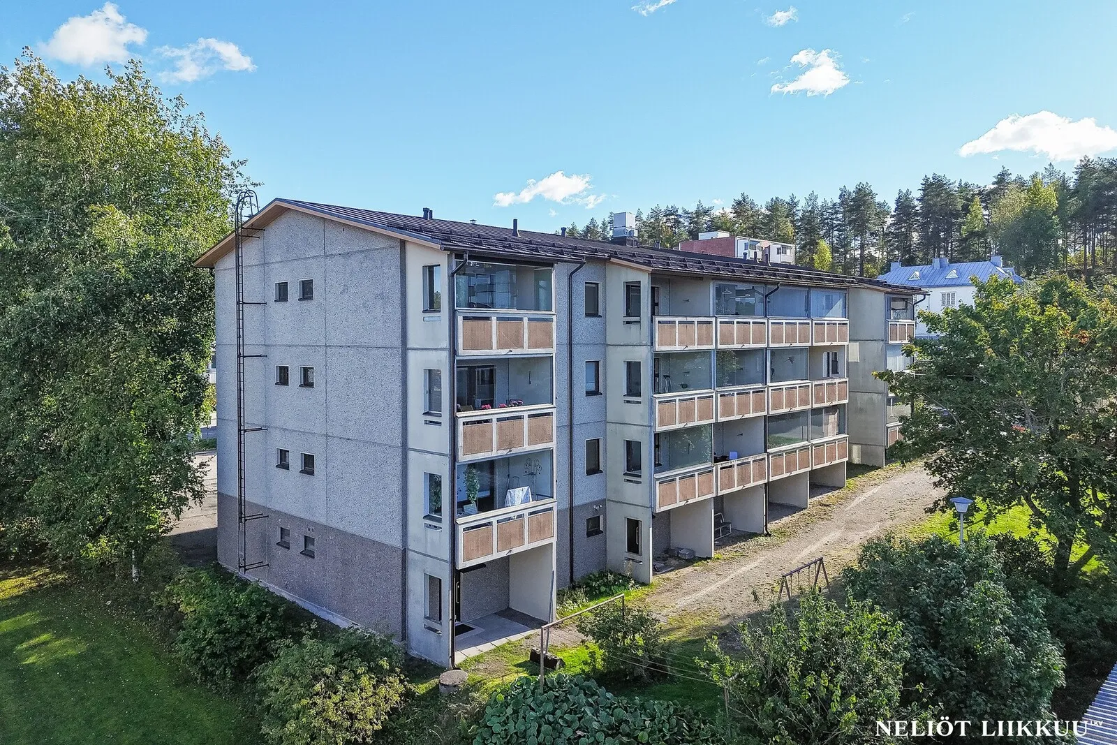 Kolmikerroksinen kerrostalo ja sen parvekkeet vihreän pihan ympäröimänä aurinkoisena päivänä.