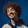 Smiling young man with curly hair wearing a blue jacket against a blue background.