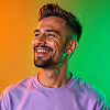 Smiling young man with beard and short hair wearing a purple shirt against a green-orange background.