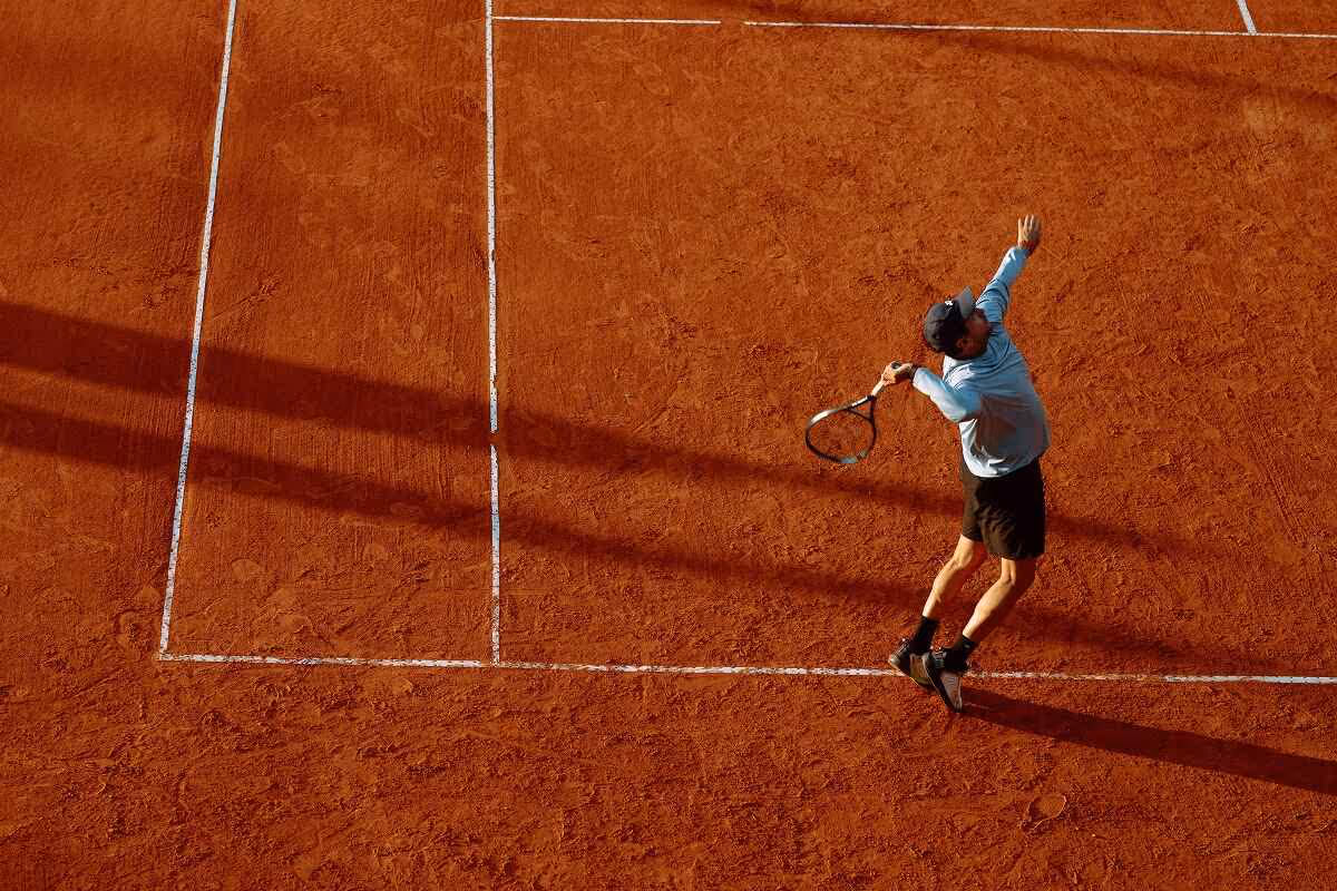 Tennis player in black shorts and blue shirt serving on a clay court with visible shadows.