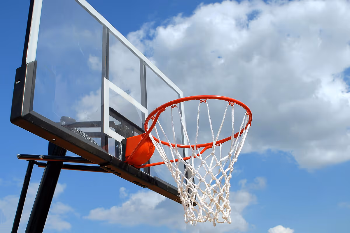 Basketball hoop and net mounted on a transparent backboard against a blue sky with some clouds.