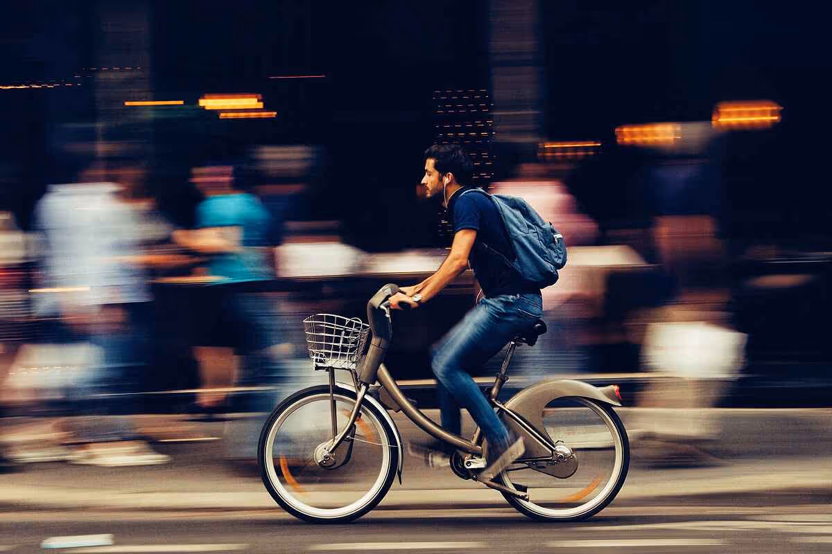 Man with backpack riding a bicycle on a city street with blurred pedestrians in the background.