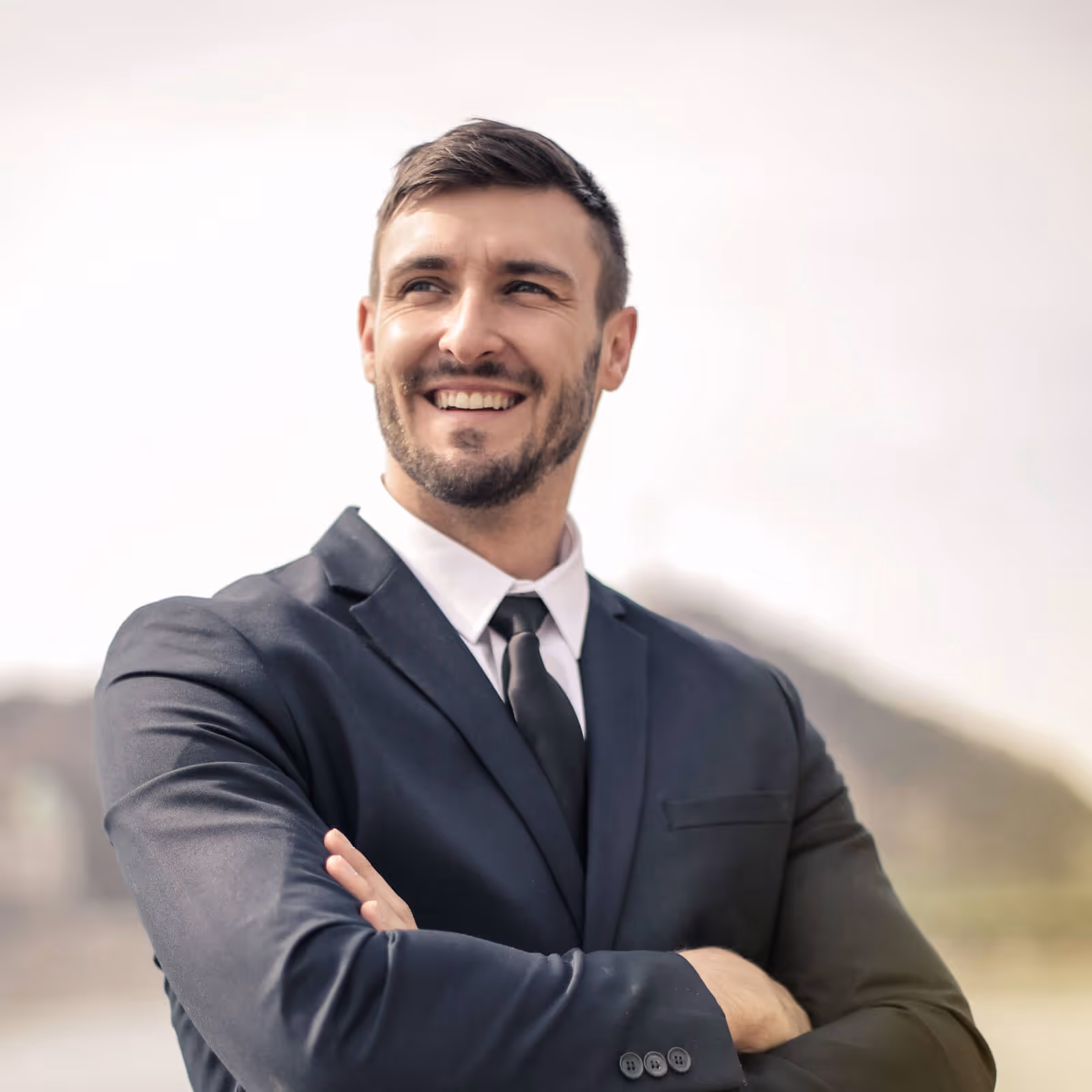 Smiling confident man in dark suit with arms crossed, looking off to the side outdoors.