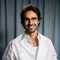 Smiling man with glasses, dark curly hair, and a white shirt against a gray curtain background.