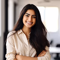 Smiling woman with long dark hair wearing a beige shirt standing with arms crossed indoors.