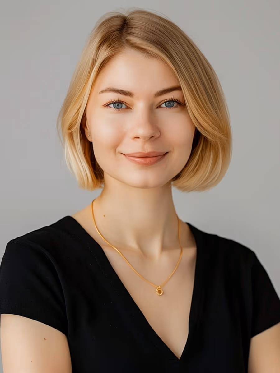 Smiling blonde woman with short hair wearing a black top and gold necklace against gray background.