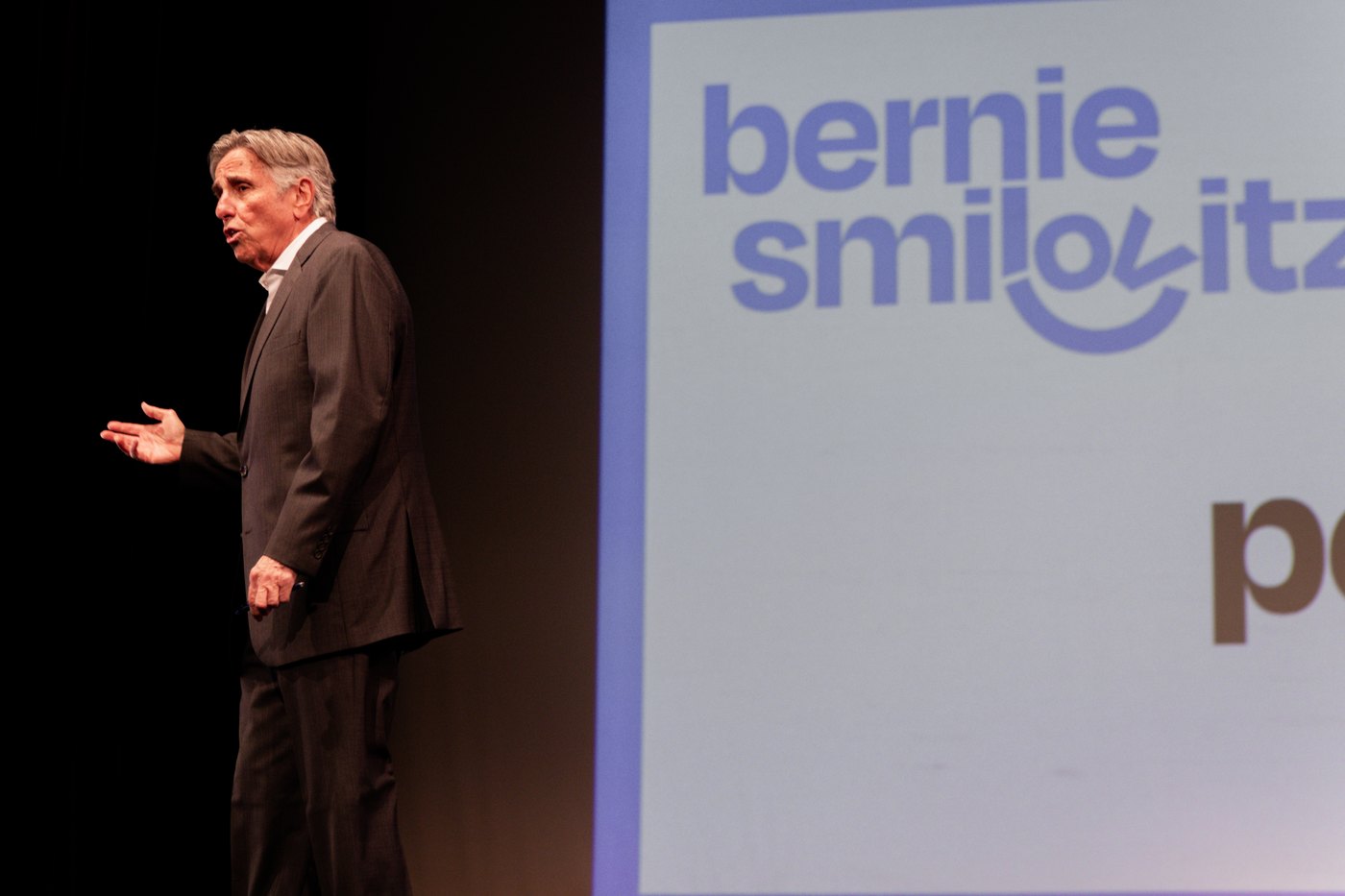 Bernie Smilovitz speaking in profile on stage beside his large logo slide