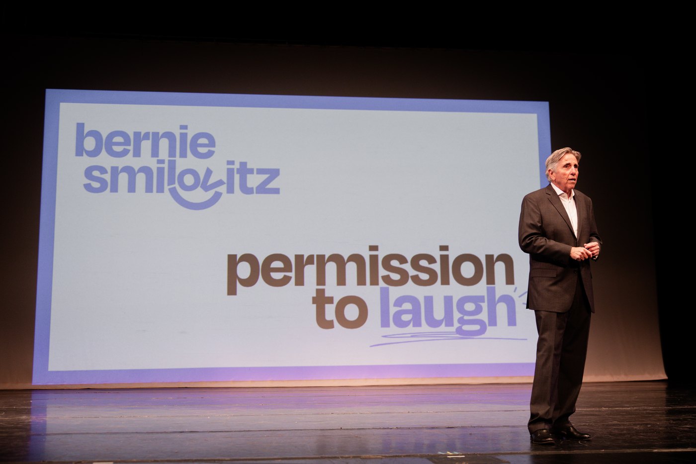 Bernie Smilovitz speaking on stage beside his Permission to Laugh keynote slide