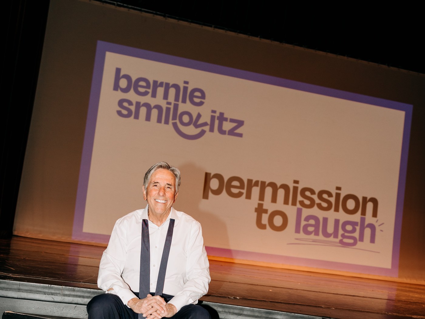 Bernie Smilovitz smiling on stage steps in shirt and suspenders before his Permission to Laugh slide