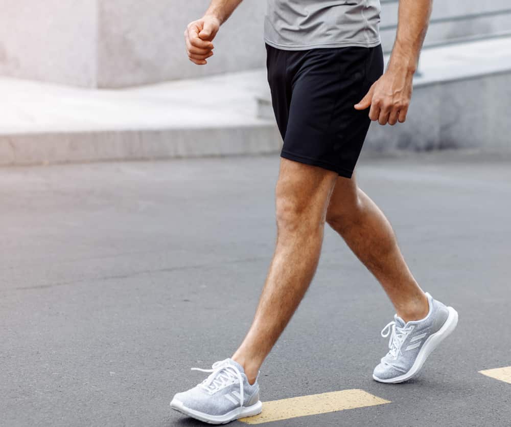 A person wearing black athletic shorts, a gray t-shirt, and gray Adidas running sneakers walking on an outdoor paved surface, captured from the waist down, suggesting light exercise or post-surgery recovery walking.
