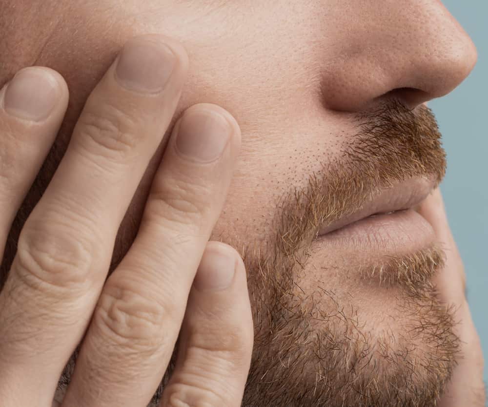 Close-up of a man with a reddish-brown stubble beard pressing his hand against his cheek, suggesting jaw or facial pain or discomfort.