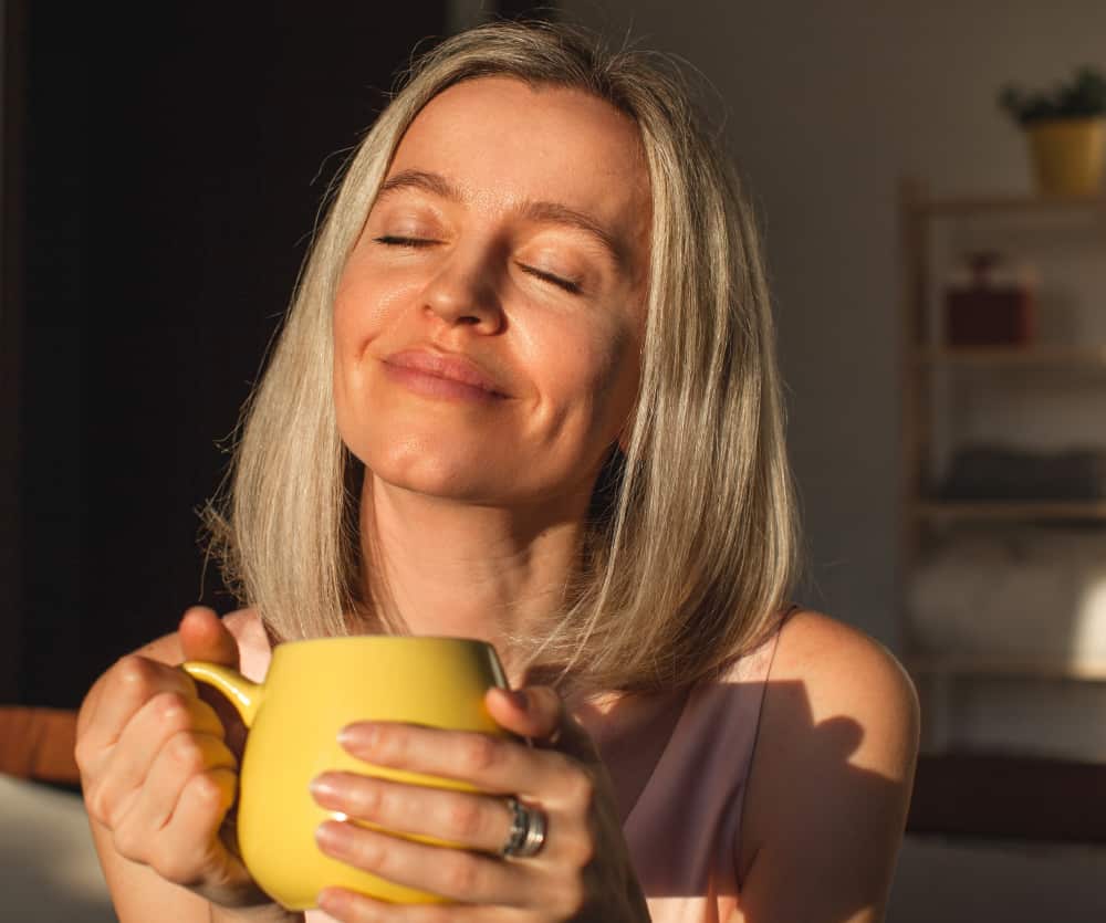 A middle-aged woman with shoulder-length gray-blonde hair smiling contentedly with her eyes closed, holding a yellow ceramic mug with both hands, bathed in warm sunlight, with a softly blurred home interior in the background.