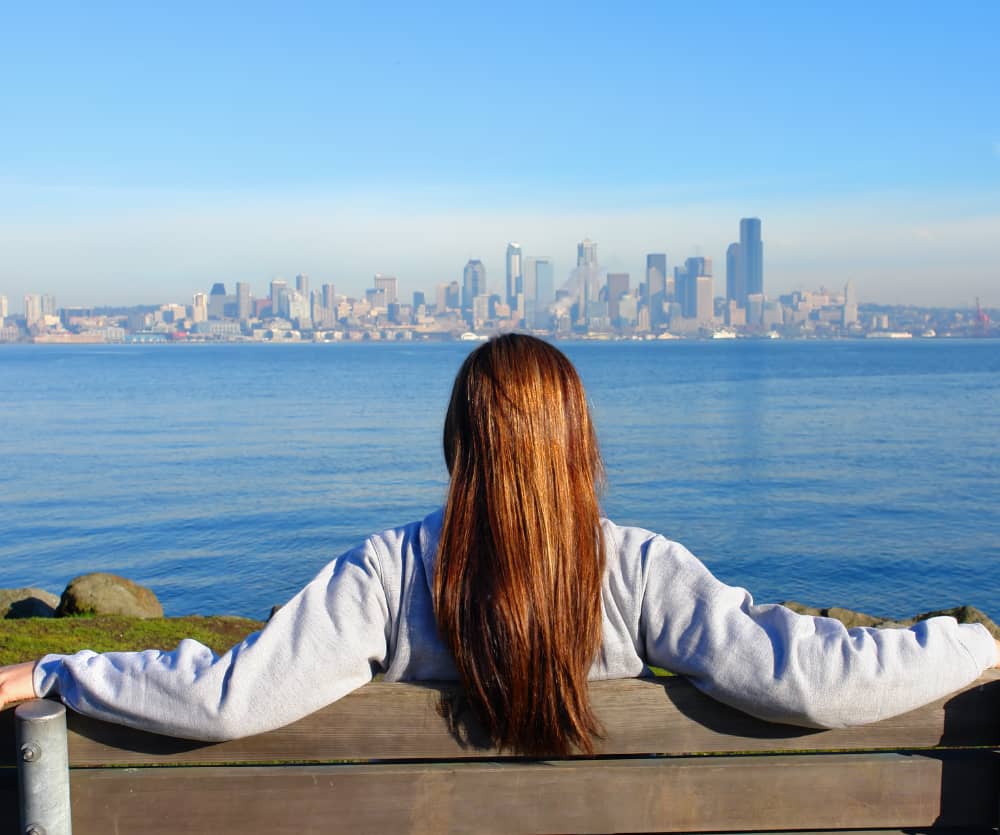 A woman with long brown hair, wearing a gray hoodie, sits on a wooden bench with her arms spread wide, facing away from the camera toward a calm blue bay and a city skyline under a clear blue sky.