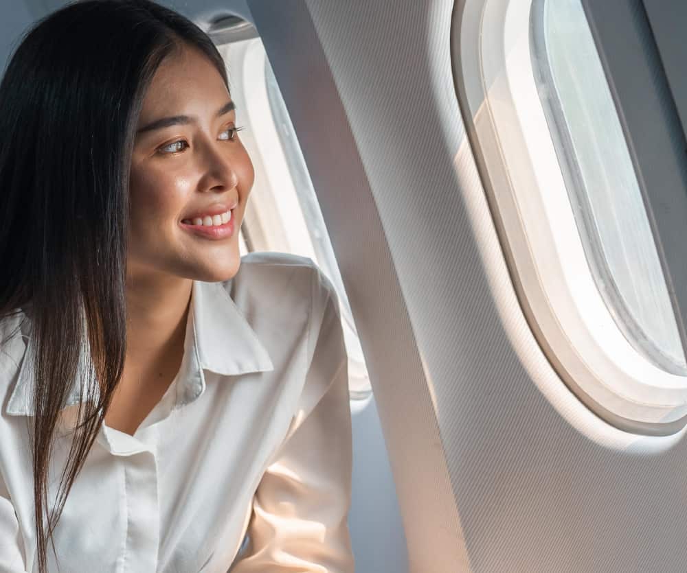 A smiling young woman with long dark hair, wearing a white button-up shirt, seated inside an airplane cabin and gazing out of the oval airplane window with natural light coming through.