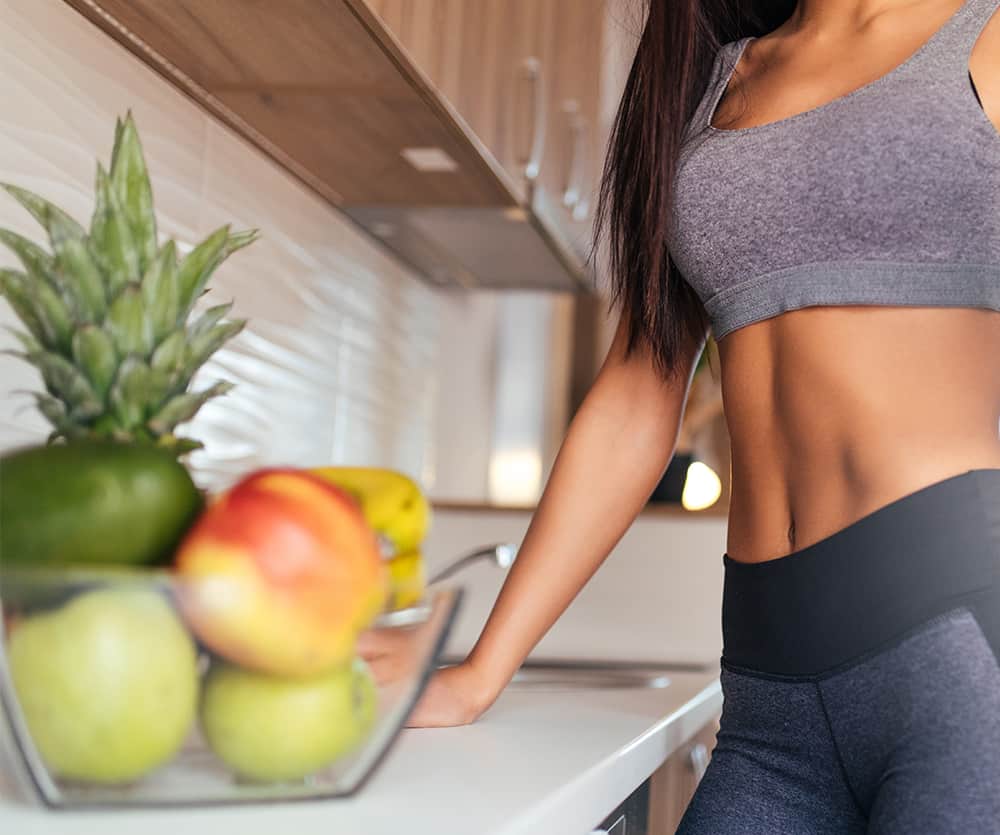 A fit woman wearing a gray sports bra and black leggings standing in a modern kitchen next to a counter with a bowl of fresh fruits including a pineapple, apples, and bananas, showcasing a healthy lifestyle.