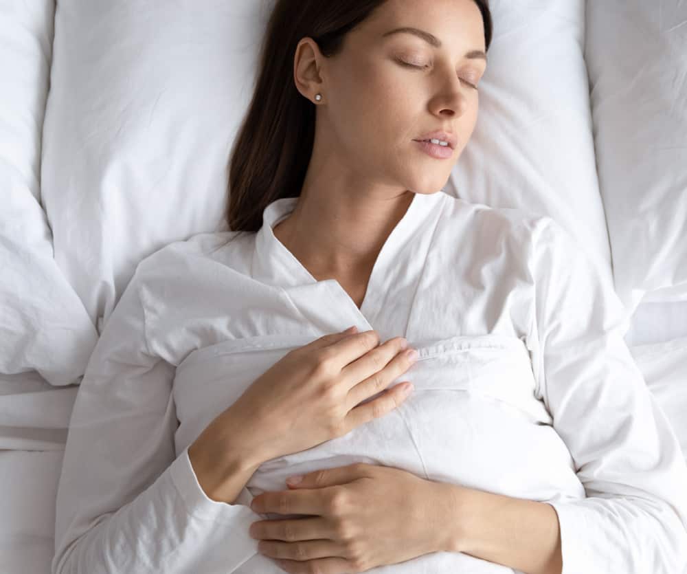 A woman lying on her back in bed on white pillows and sheets, wearing a white robe, with her eyes closed and hands resting on her abdomen, illustrating a comfortable sleeping position after a tummy tuck surgery.