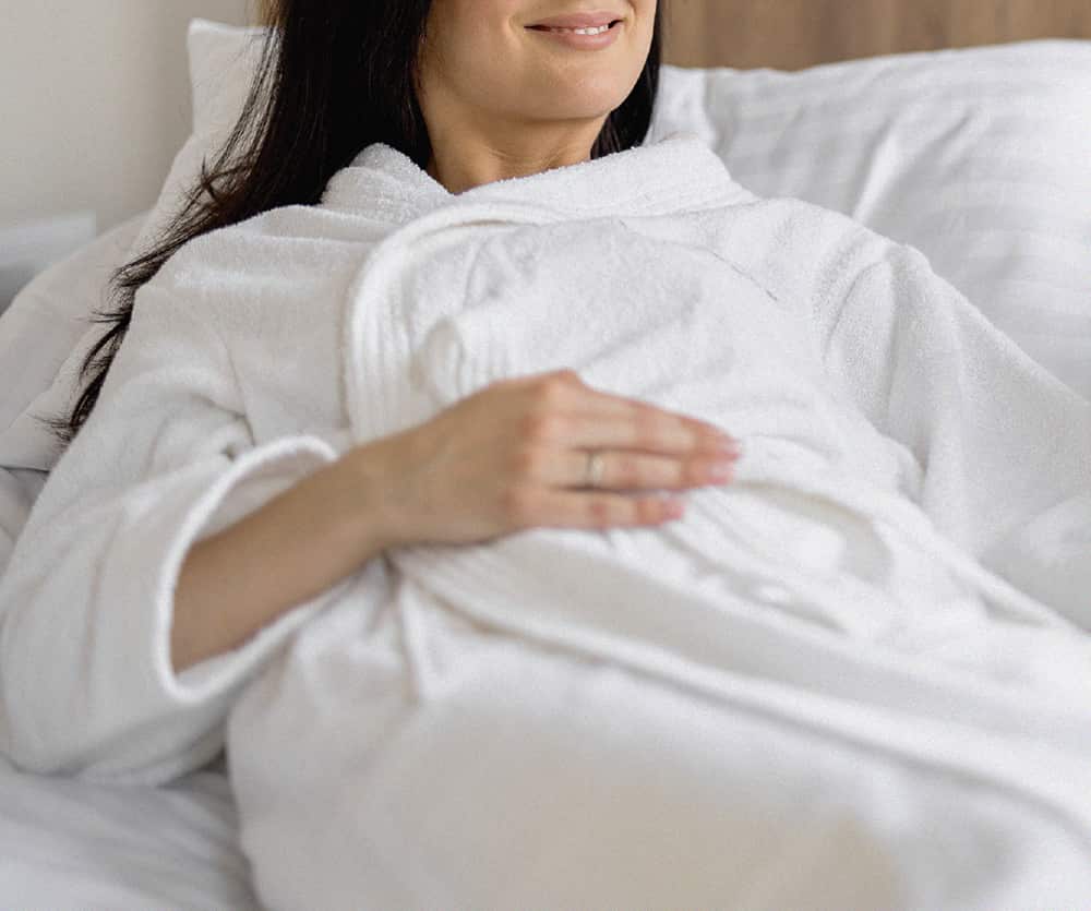 A smiling dark-haired woman wearing a white fluffy bathrobe resting in bed with white pillows and bedding, gently resting her hand with a ring on her chest, illustrating how to sleep comfortably after breast augmentation surgery.