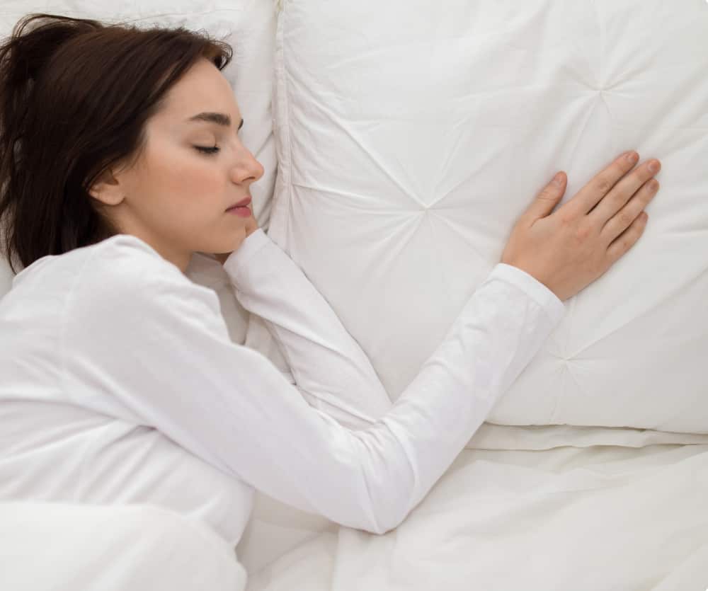 A young woman with dark hair sleeping peacefully on her side in bed, wearing a white long-sleeve top, resting her head on a white pillow and hugging it with both arms, covered by a white duvet.