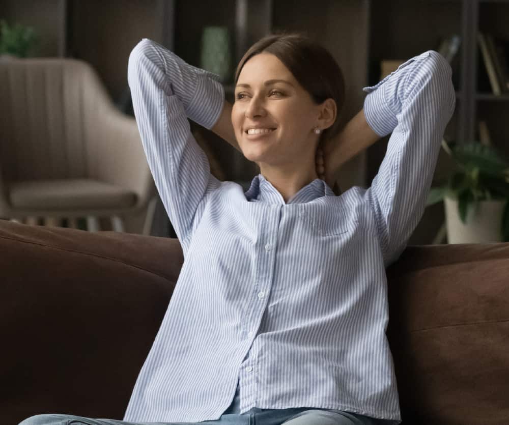 A young woman with brown hair pulled back, wearing a light blue striped button-up shirt and jeans, sitting relaxed on a brown sofa with her hands clasped behind her head, smiling contentedly in a cozy living room setting.