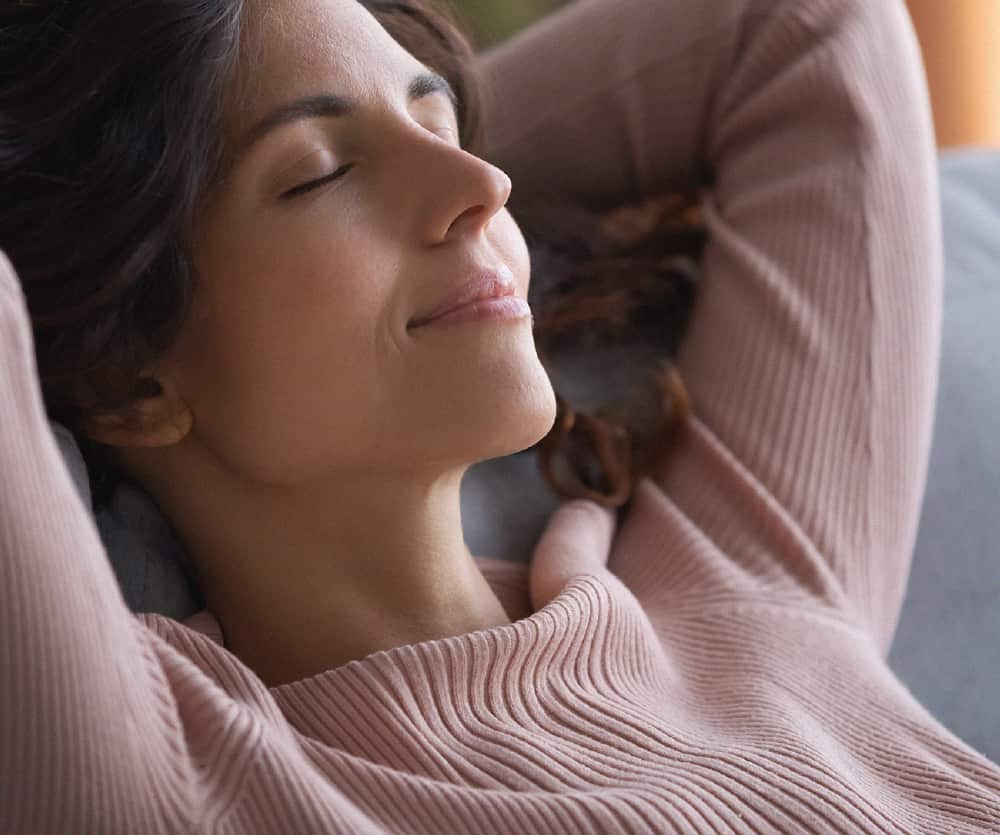 A relaxed woman with dark hair wearing a pink ribbed sweater, reclining on a sofa with her eyes closed and a calm smile, hands clasped behind her head, conveying comfort and peaceful recovery.