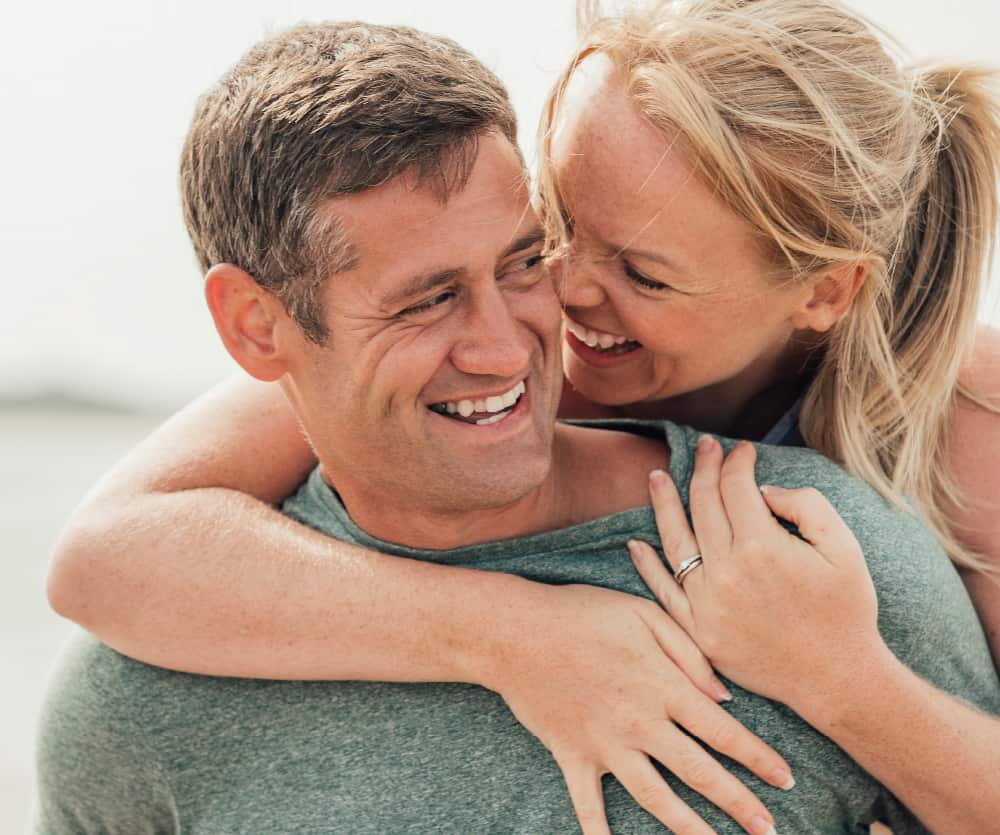 A smiling couple sharing a joyful moment outdoors, with a woman wrapping her arms around a man's shoulders from behind, both laughing and looking at each other. The woman wears a ring on her finger.