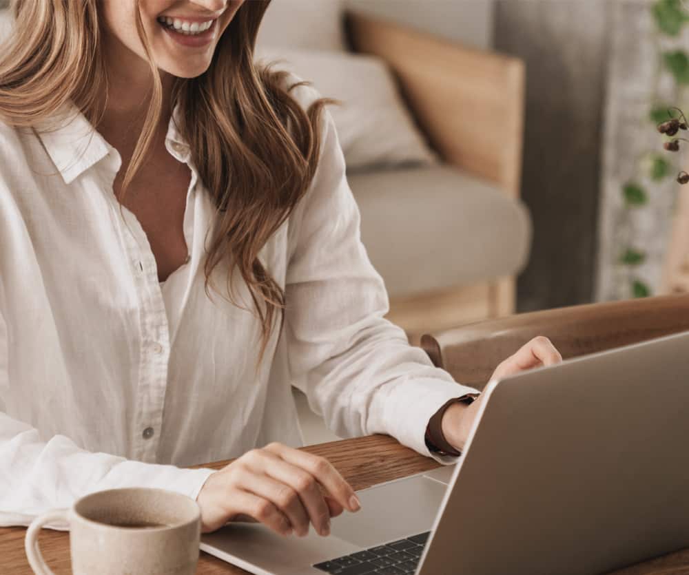 A smiling woman with long blonde hair, wearing a white linen shirt and a brown watch, typing on a laptop at a wooden desk with a beige ceramic coffee mug nearby, in a cozy home setting with soft furnishings and a green plant in the background.