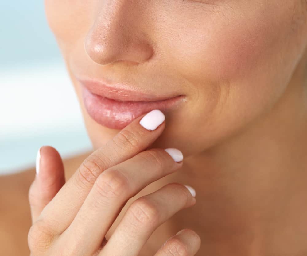 Close-up of a woman's lower face with naturally glossy lips, gently touching her mouth with her fingers, showcasing well-manicured nails with white polish, in the context of what to expect after lip fillers.