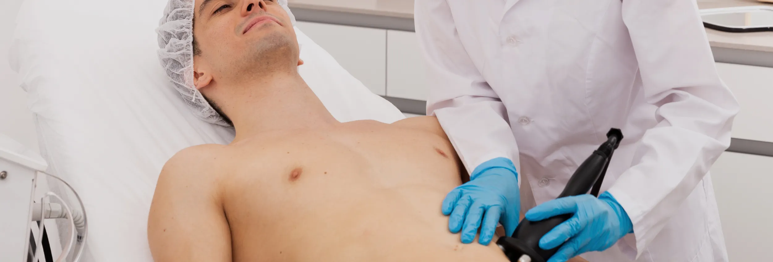 A male patient lying shirtless on a treatment bed wearing a disposable hair net, while a medical professional in a white lab coat and blue latex gloves applies a body sculpting device to his abdomen in a clinical setting.