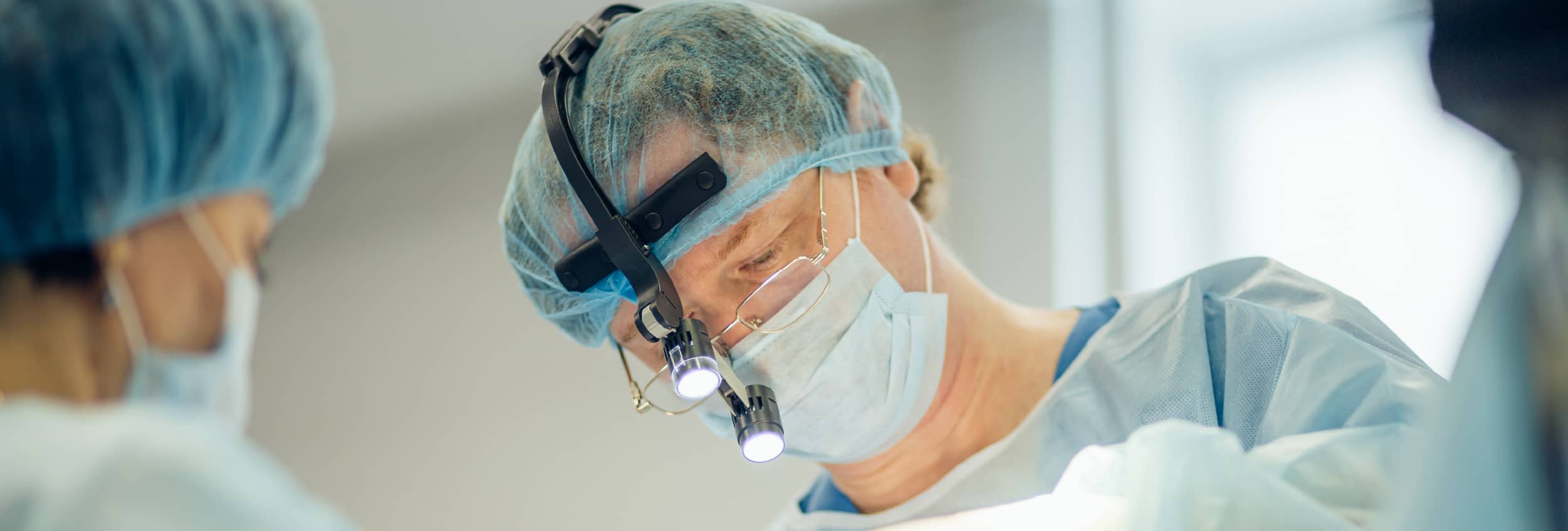 A male patient lying shirtless on a treatment bed wearing a disposable hair net, while a medical professional in a white lab coat and blue latex gloves applies a body sculpting device to his abdomen in a clinical setting.