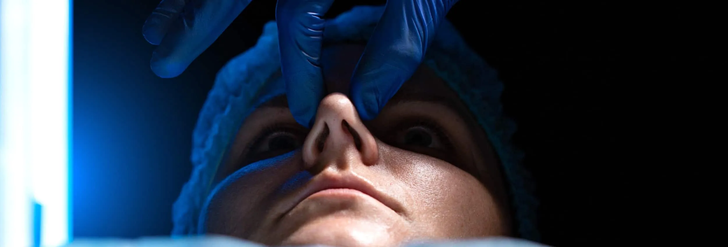 A male patient lying shirtless on a treatment bed wearing a disposable hair net, while a medical professional in a white lab coat and blue latex gloves applies a body sculpting device to his abdomen in a clinical setting.