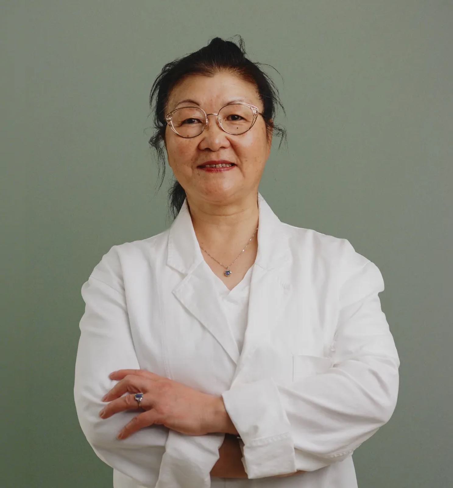 Dr. Nancy Xiaonan Du, a woman wearing a white medical lab coat, clear-framed glasses, and a delicate necklace, smiling with arms crossed in a professional portrait photo.