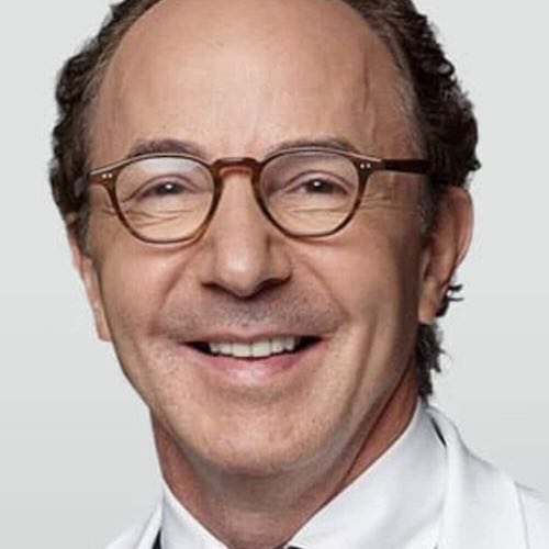 Professional headshot of a middle-aged man with short dark hair, wearing brown tortoiseshell glasses and a white medical coat, smiling warmly at the camera.
