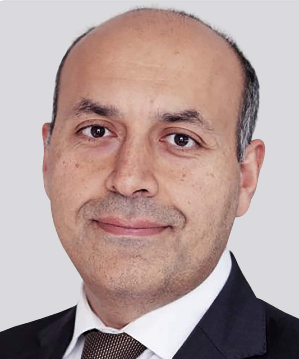 Professional headshot of Professor Ash Mosahebi, a middle-aged man with a shaved head and short dark stubble, wearing a dark suit jacket, white dress shirt, and a brown patterned tie, smiling gently at the camera.